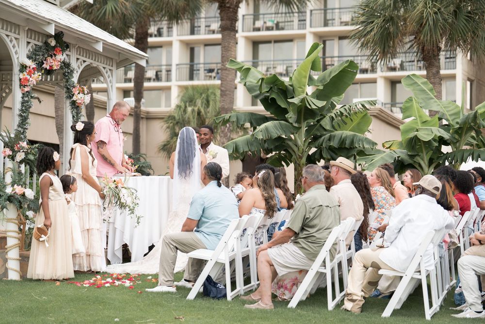 Outdoor wedding ceremony with guests seated in white chairs on a lawn beside a hotel building with tropical plants.