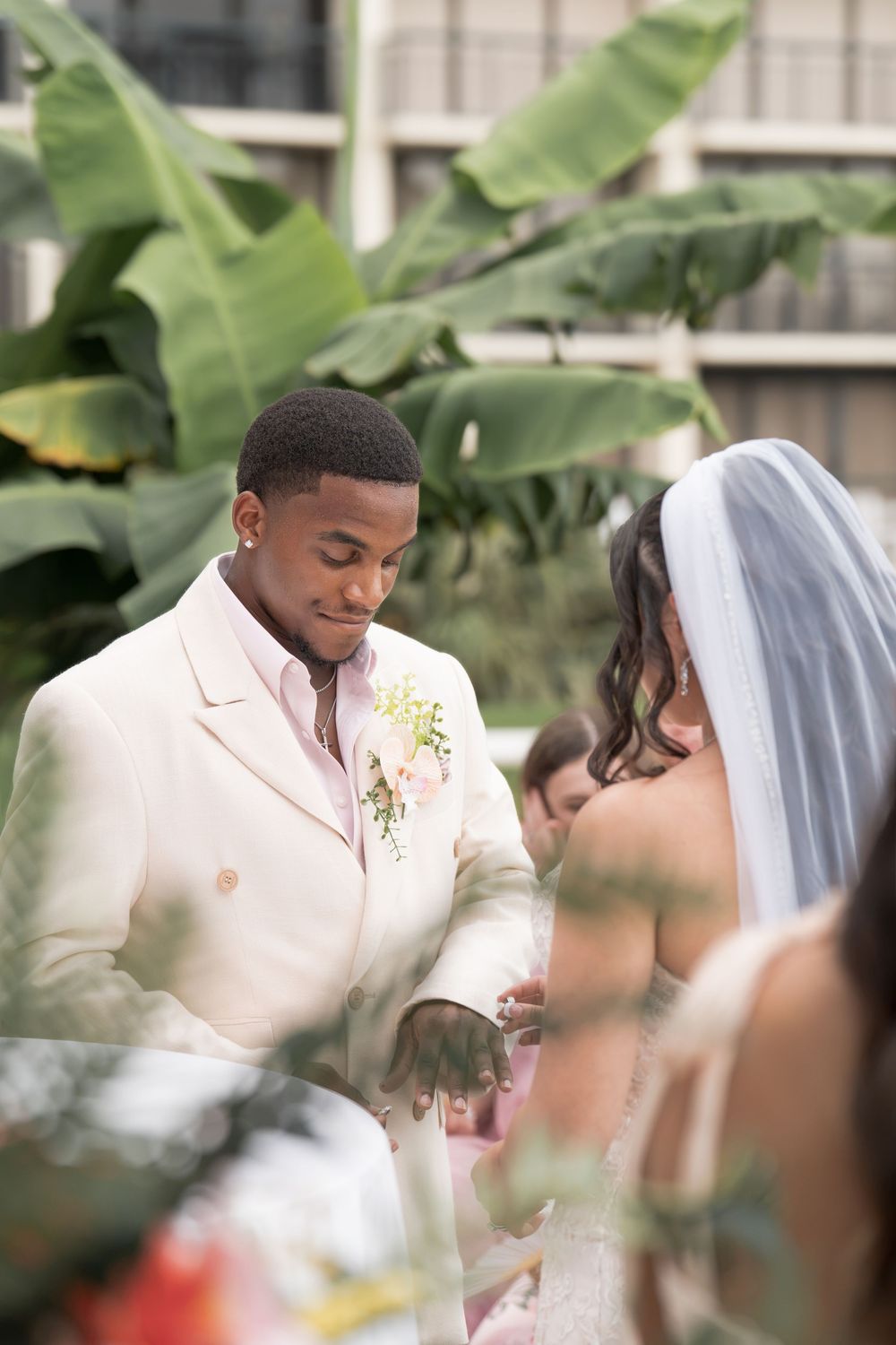 Groom in ivory suit placing wedding ring on brides hand during outdoor ceremony with tropical backdrop.