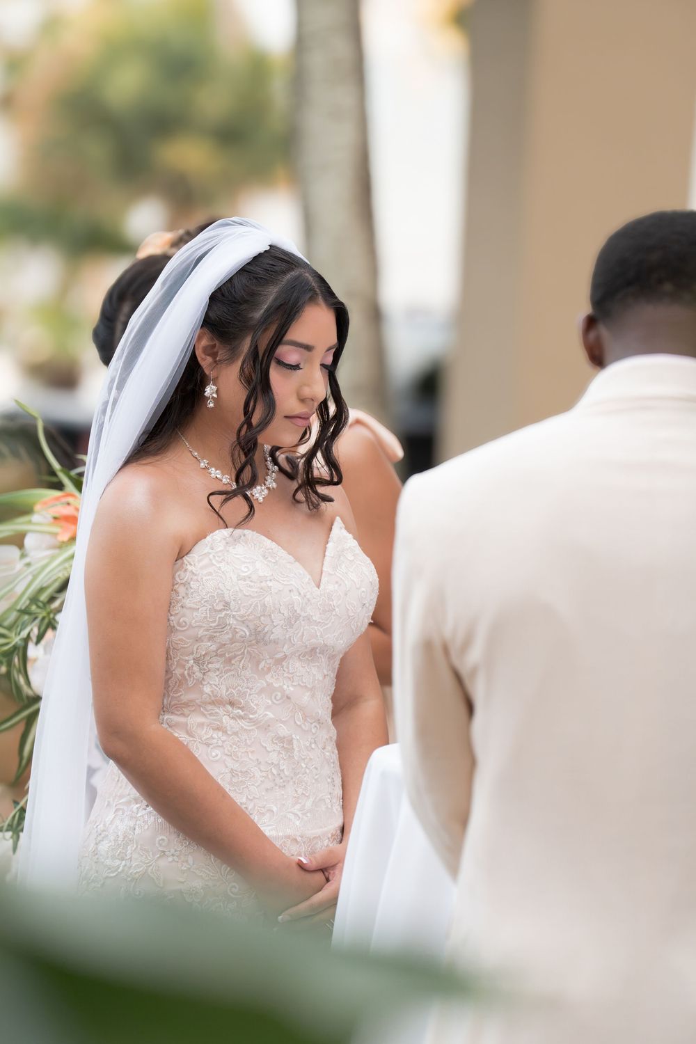 Bride in strapless lace wedding gown and veil bowing head during outdoor wedding ceremony exchange of vows.