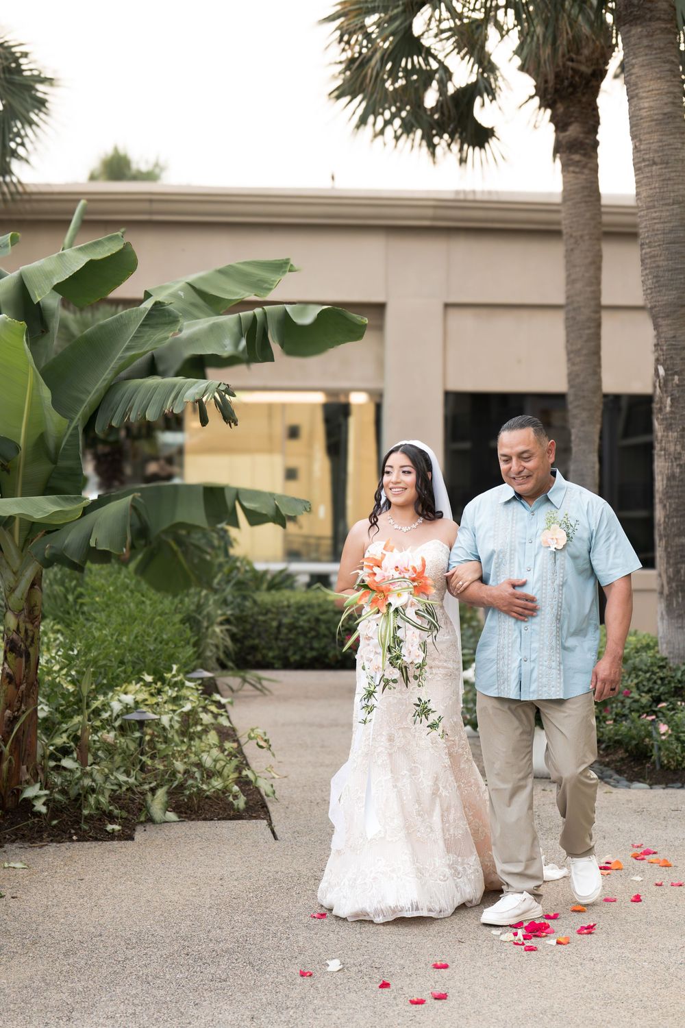 Bride in lace gown walks with escort along rose petal path surrounded by tropical palms.
