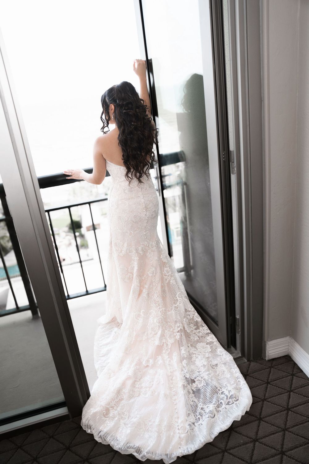 Bride in strapless lace mermaid wedding gown standing at balcony door looking out at scenic view.