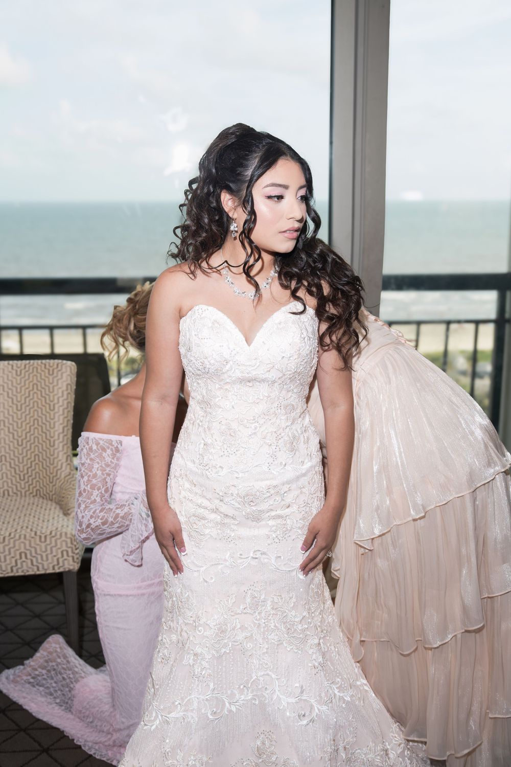 Bride in strapless lace mermaid wedding gown with curly hair poses near ocean view window.