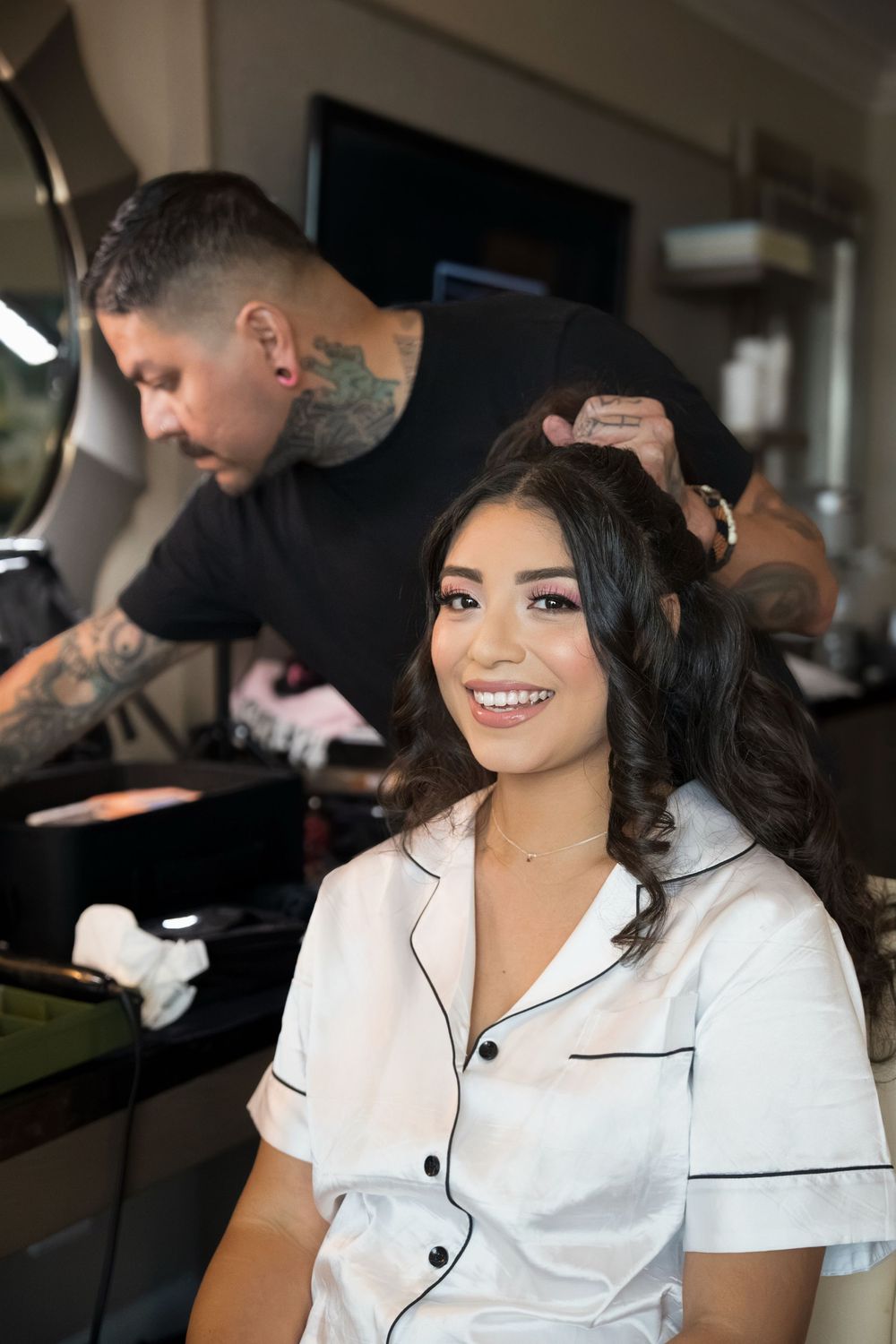 Smiling bride gets hair styled by tattooed stylist in salon before wedding.