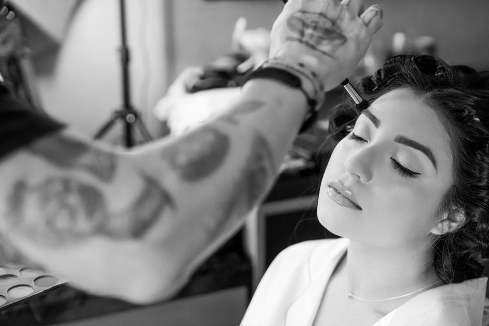 Black and white photo of a tattooed makeup artist applying makeup to a young woman with curly hair.