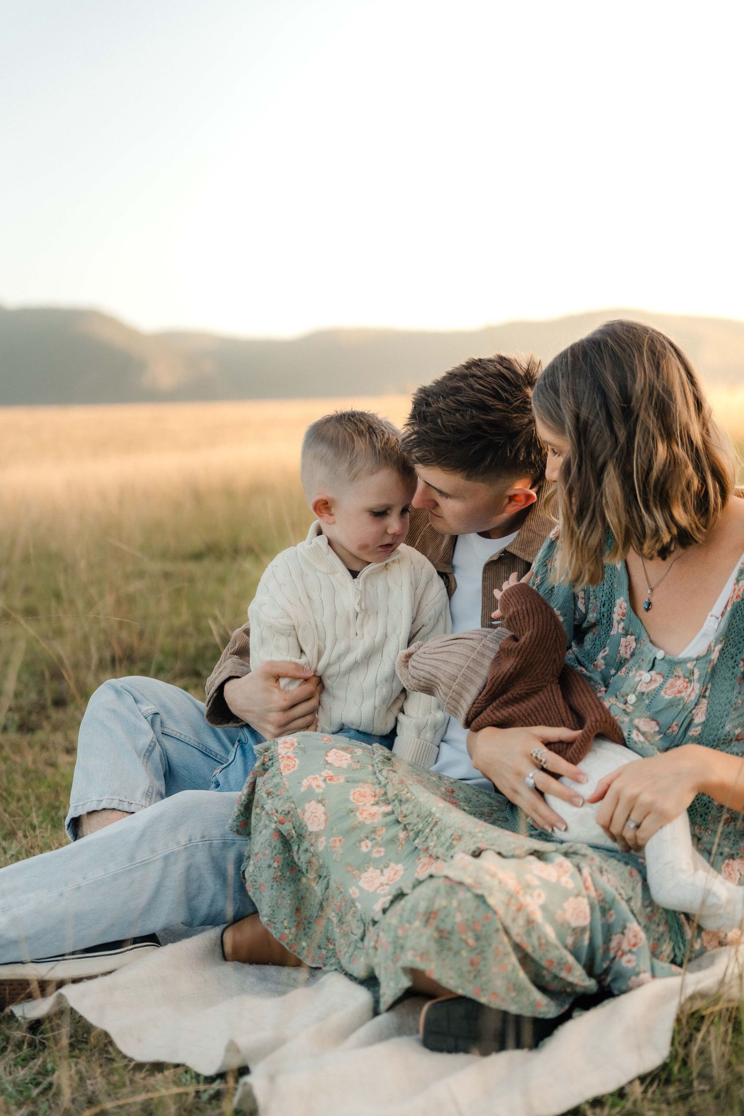 Parents with toddler and newborn baby sitting on a blanket in an open field at golden hour.