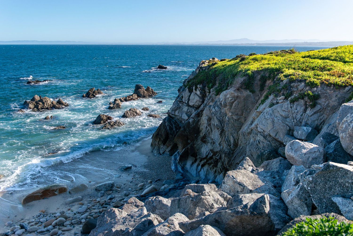Rocky coastal cliffs meet crashing ocean waves under a clear blue sky, with green vegetation along the rugged shoreline.