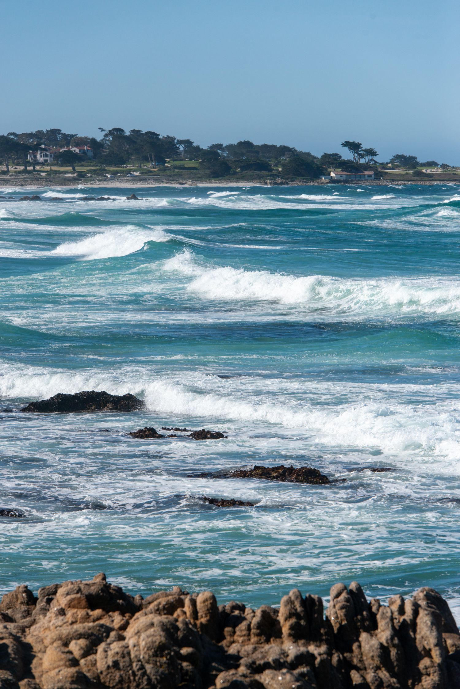 Turquoise ocean waves crash over rocky shoreline with a tree-lined coastal town visible in the background under clear blue skies.