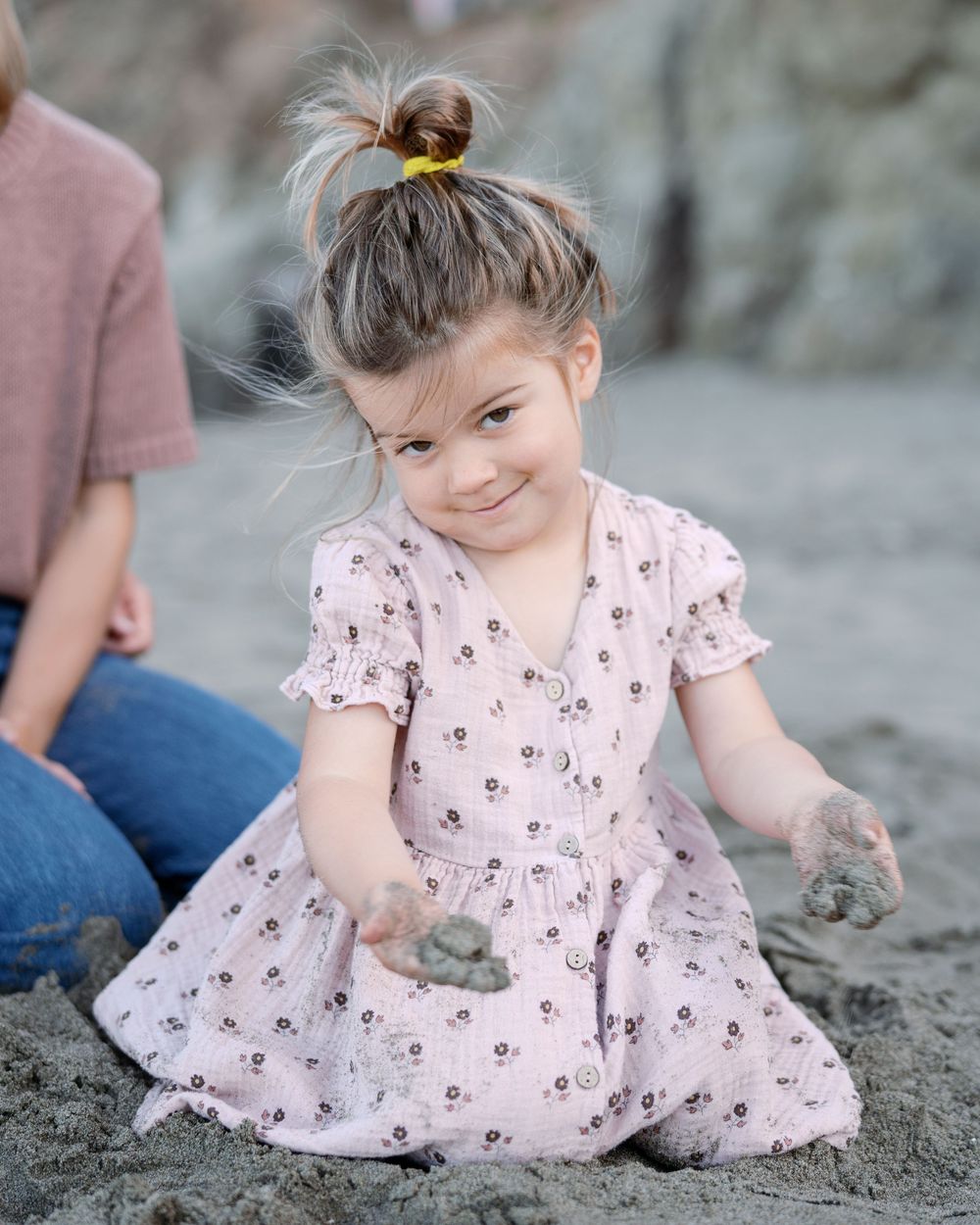 Young girl in pink floral dress playing with sand on beach, smiling cutely with hair in bun.