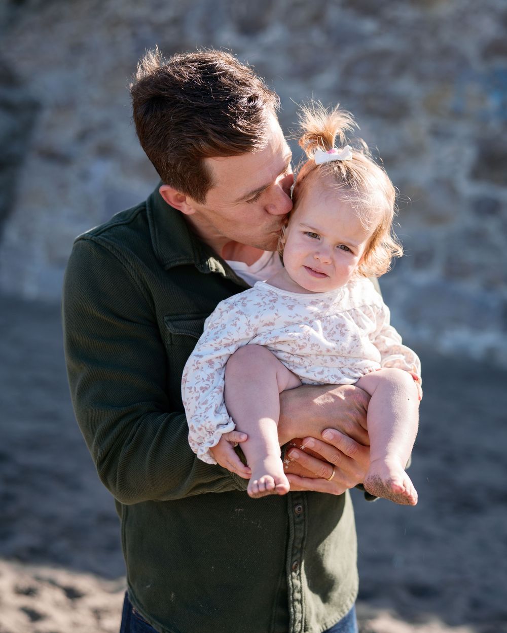 A father tenderly kisses his baby girl while holding her in his arms outdoors.