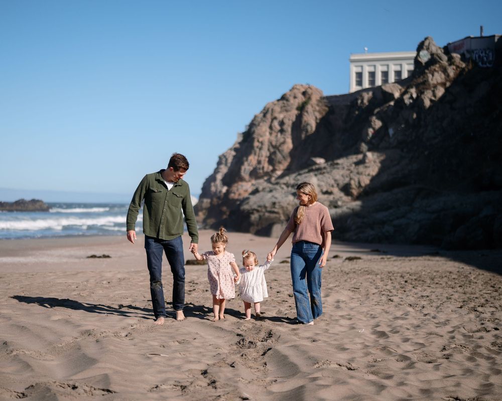 Family of four walking hand-in-hand on a sandy beach with rocky cliffs and blue sky backdrop.