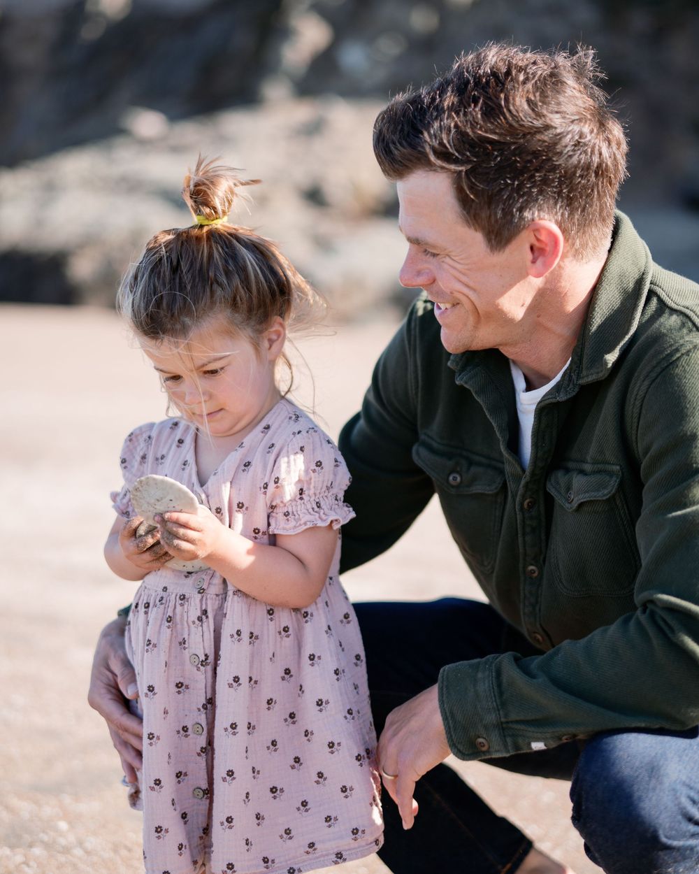 A smiling father crouches beside his young daughter in a floral dress as she examines a rock on the beach.