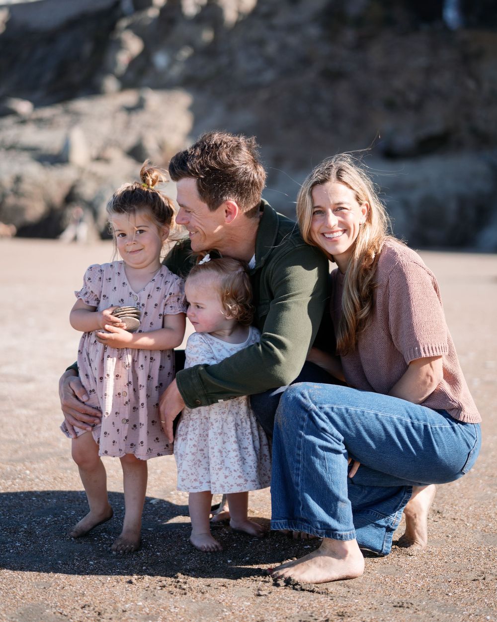 Family of four poses together on a sandy beach, parents smiling with two young daughters in floral dresses.