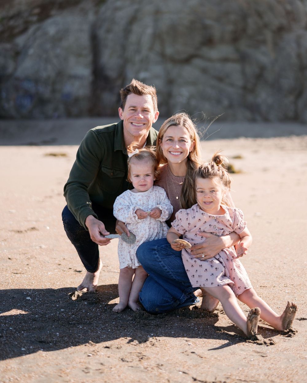 Happy family of four smiling together on a sandy beach, with rocky cliffs in the background on a sunny day.