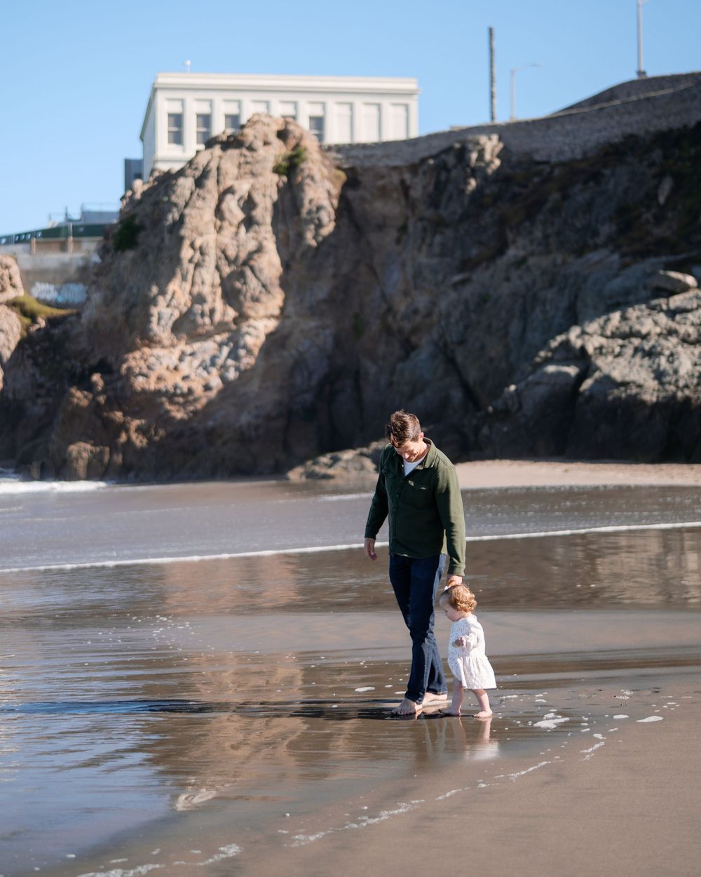 A mother and toddler walk hand-in-hand on a sandy beach with rocky cliffs and a white building in the background.