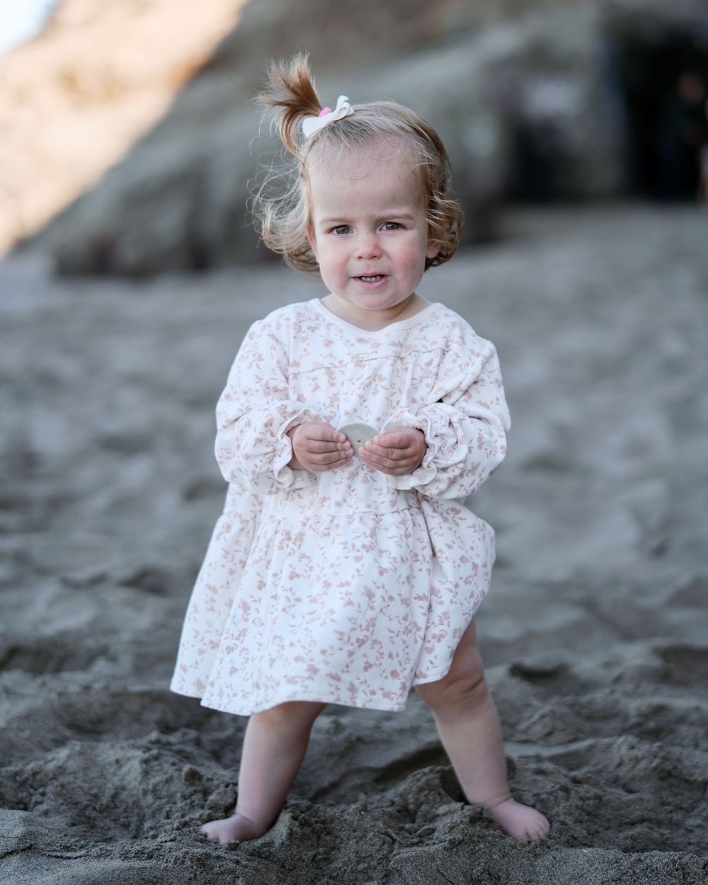 An adorable baby girl in a white floral dress stands barefoot on sandy beach.