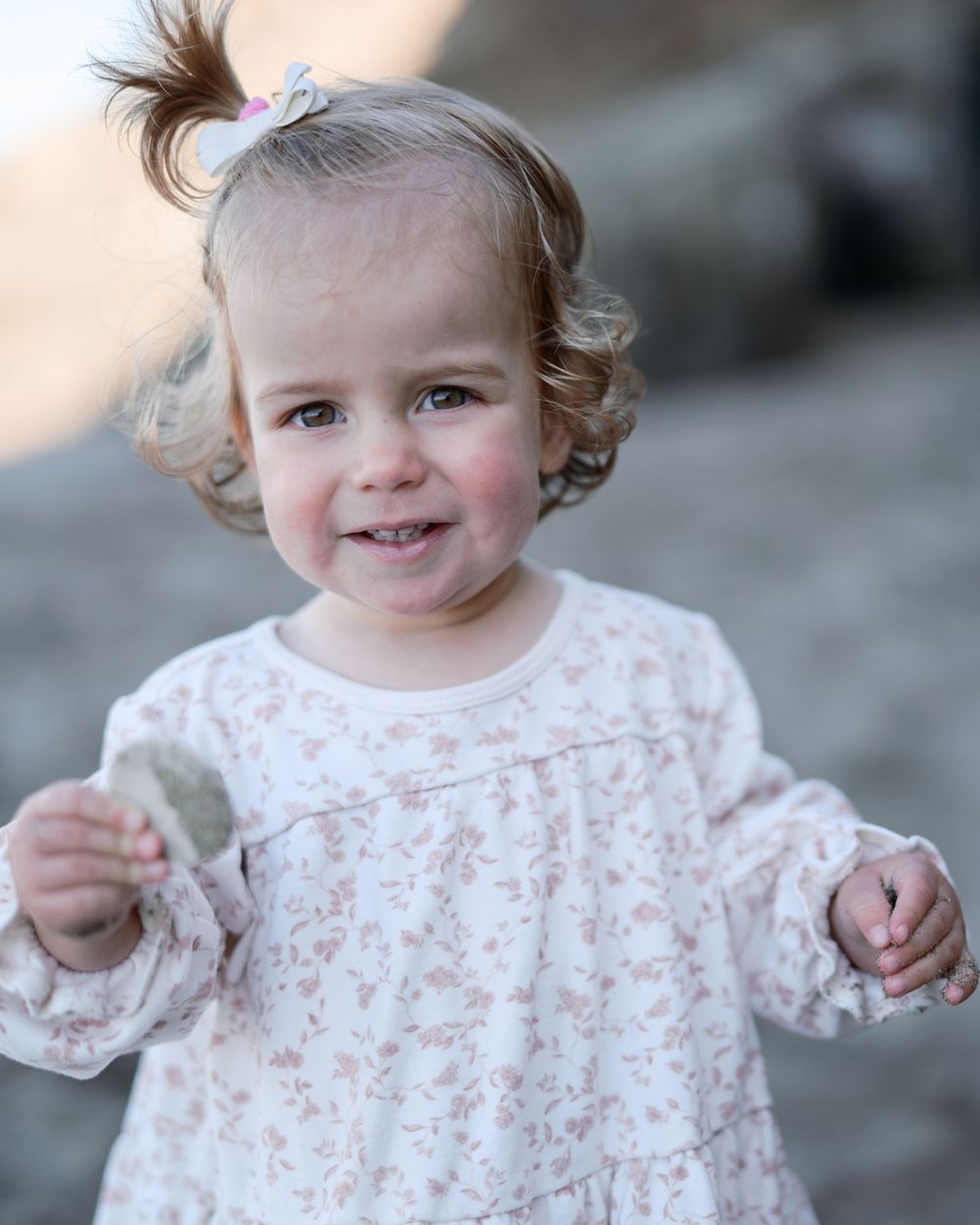 Adorable toddler girl in a floral dress smiles while holding a small object outdoors.