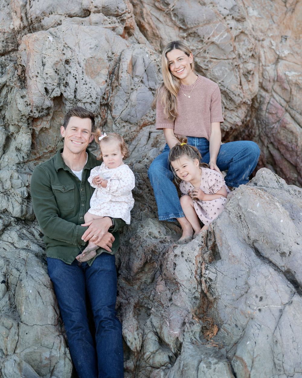 Happy family of four posing on rocky beach, parents holding two young daughters in casual outfits.