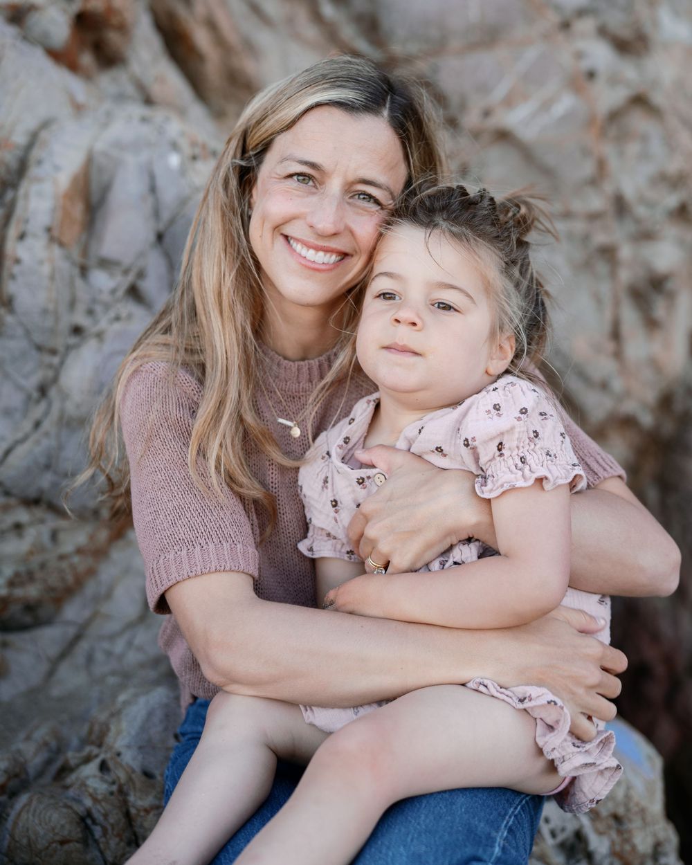 Smiling woman with long hair hugging a young girl in pink dress, sitting on rocks outdoors.