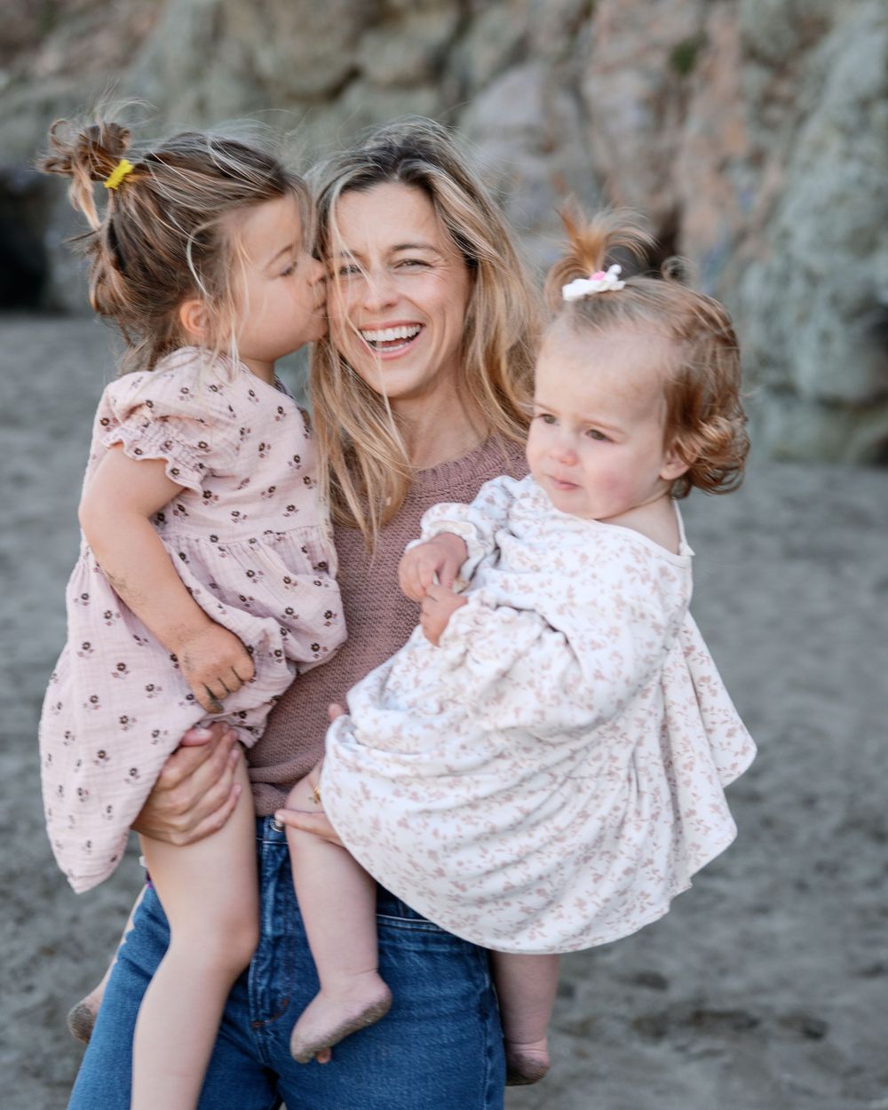 A smiling woman holds two young girls in floral dresses on a beach, one child giving her a sweet kiss.