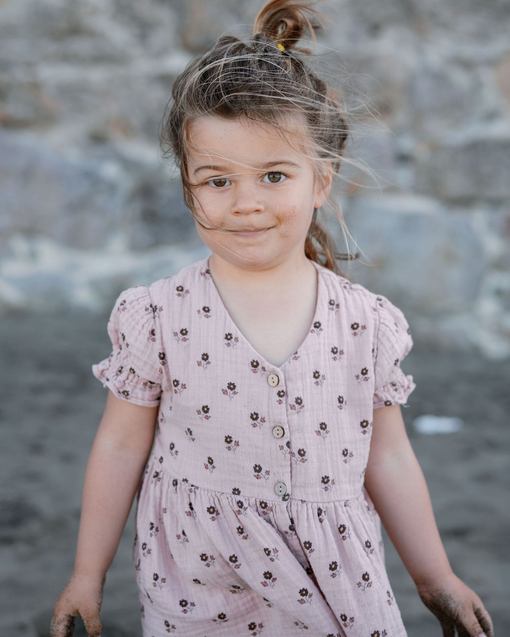 Sweet young girl in pink floral dress with hair up smiles softly in front of a stone wall backdrop.