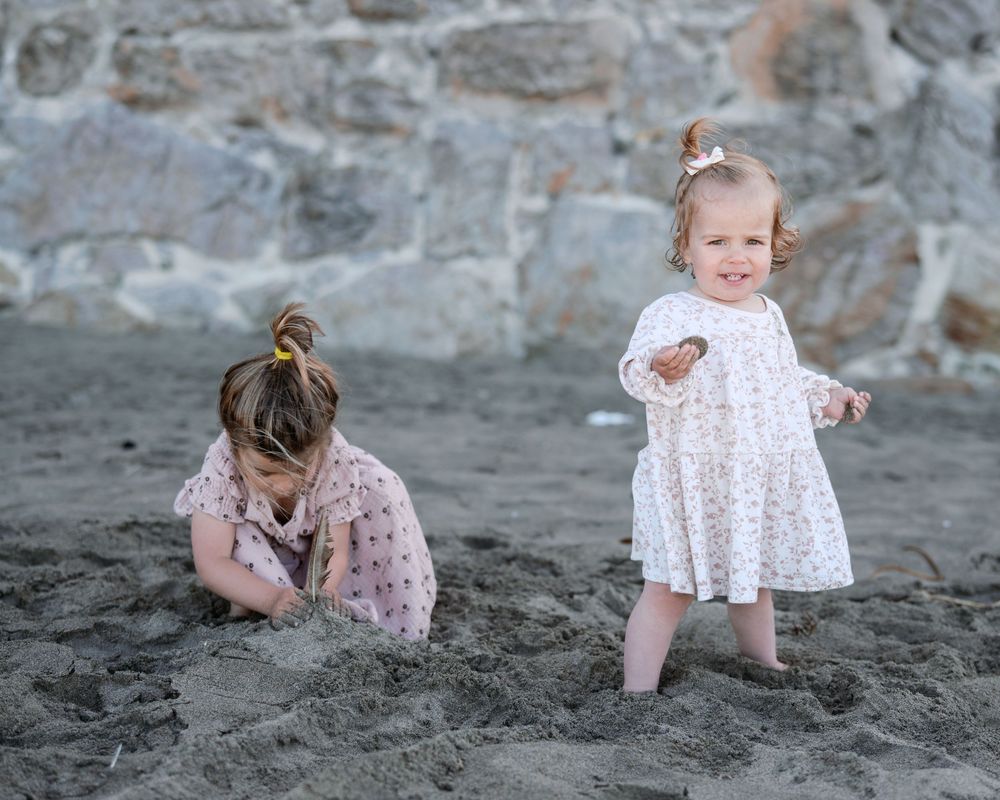Two young girls in floral dresses playing in sand on a beach near a stone wall.