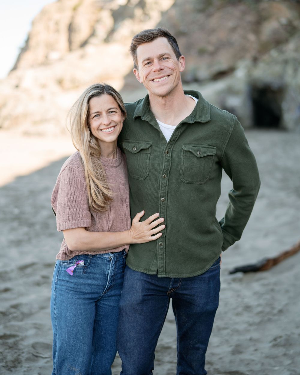 A smiling couple poses together on a sandy beach, the woman in pink and man in a green shirt.