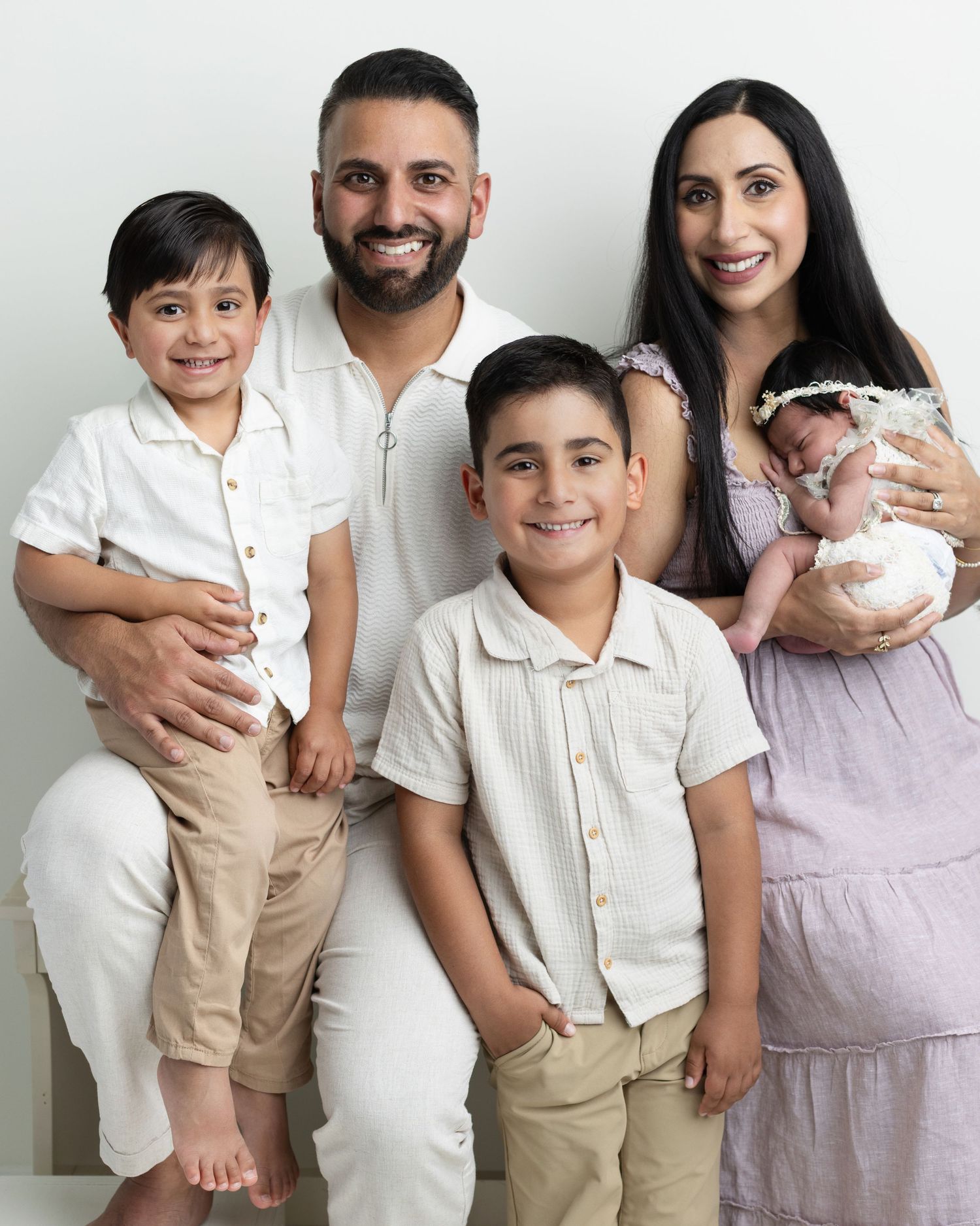Happy family of five posing together, parents holding young children and a newborn baby, wearing light casual outfits.