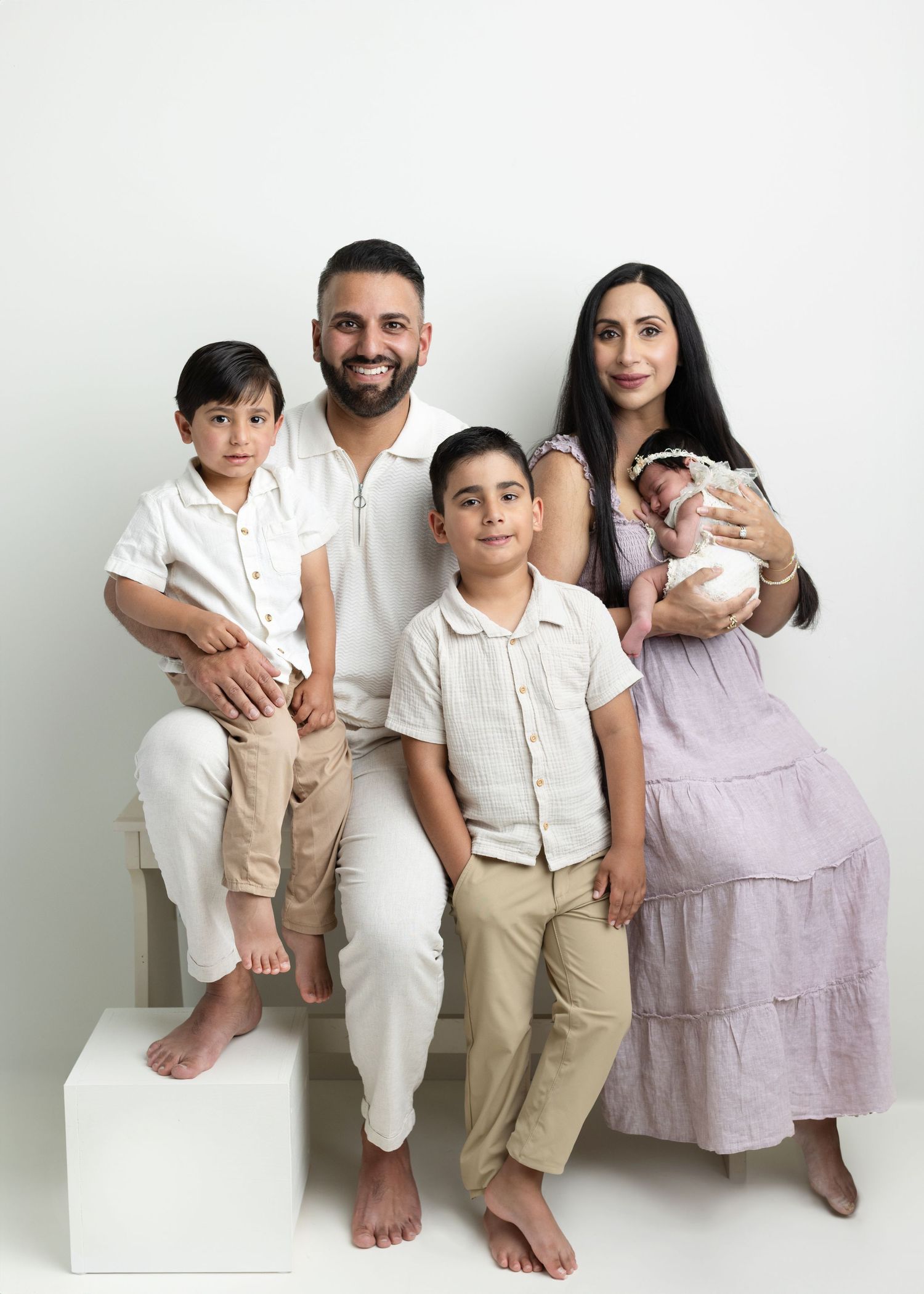 Family of five posing for a newborn portrait session, parents with two young boys and a swaddled baby, against a white background.