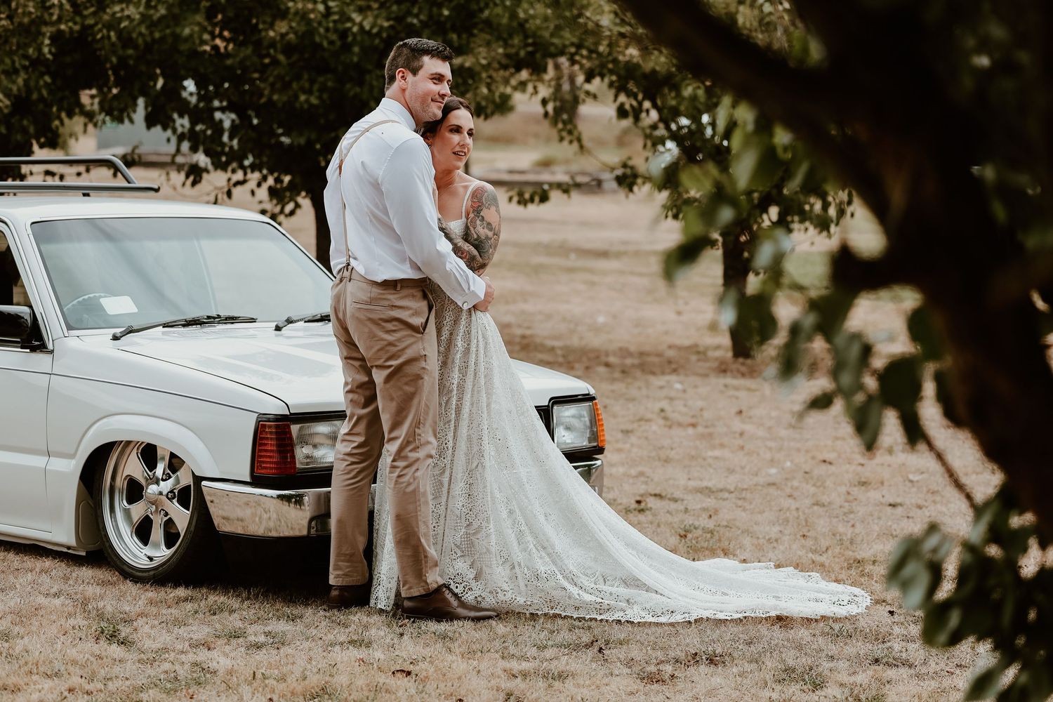 Bride and groom posing together in front of a vintage white car in a rustic outdoor setting.