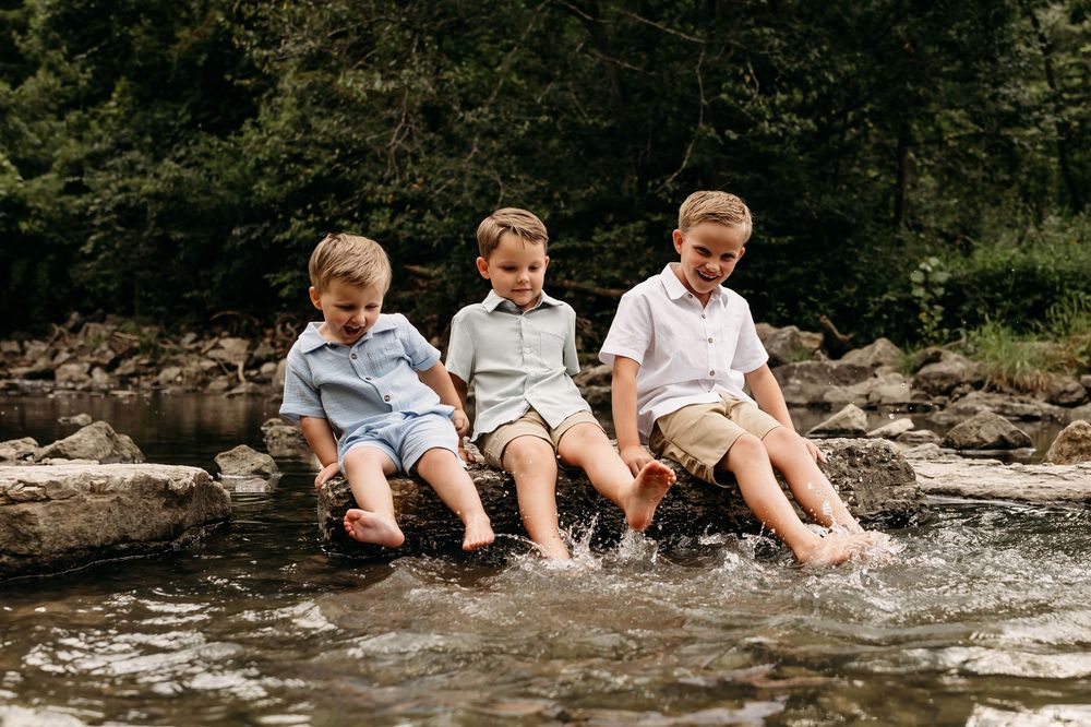 Three young boys sitting on rocks splashing their feet in a shallow creek surrounded by lush green trees.