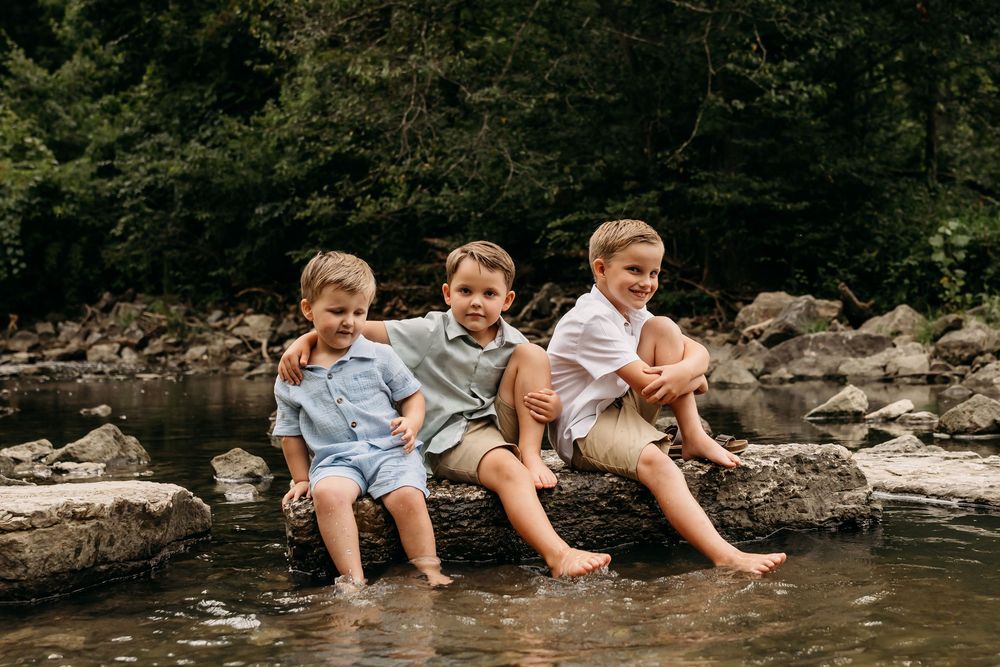 Three young boys sitting on rocks by a creek, smiling, with bare feet in the water.