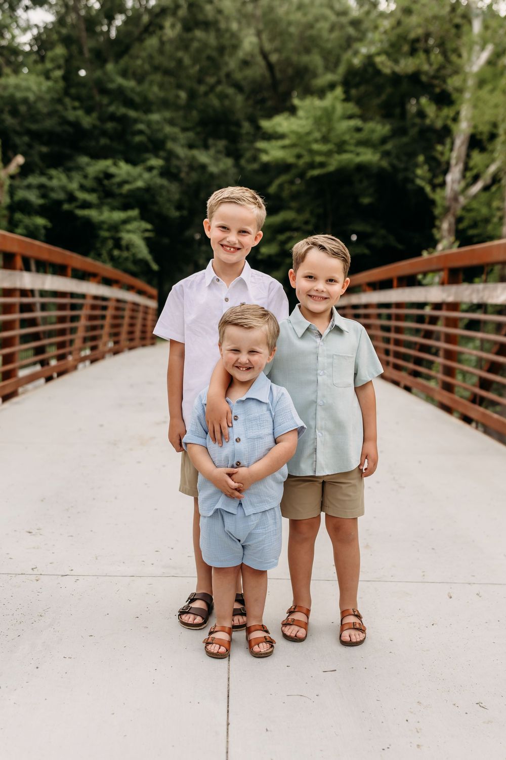 Three young boys in coordinating blue and khaki outfits pose together on a bridge surrounded by lush green trees.