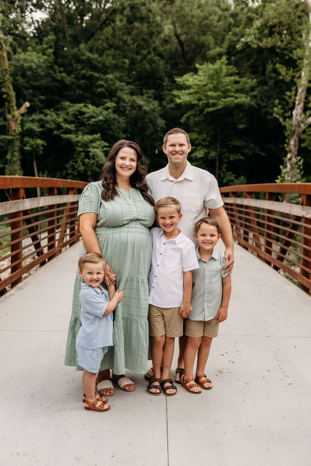 A happy family of five poses on a wooden bridge surrounded by lush green trees, with a pregnant mother in a sage dress.