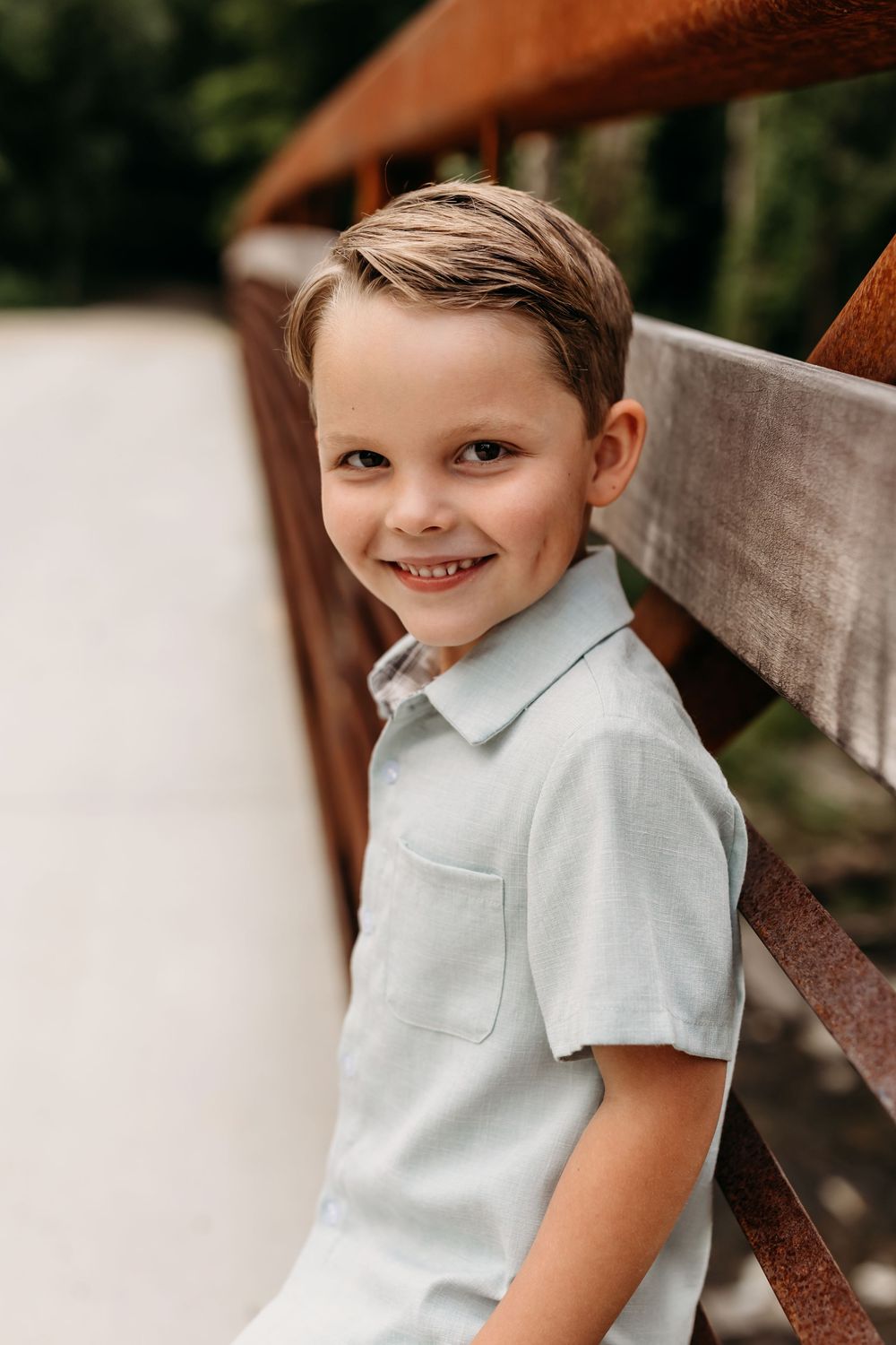 Smiling young boy in a light blue polo shirt leaning against a rustic wooden bridge railing outdoors.