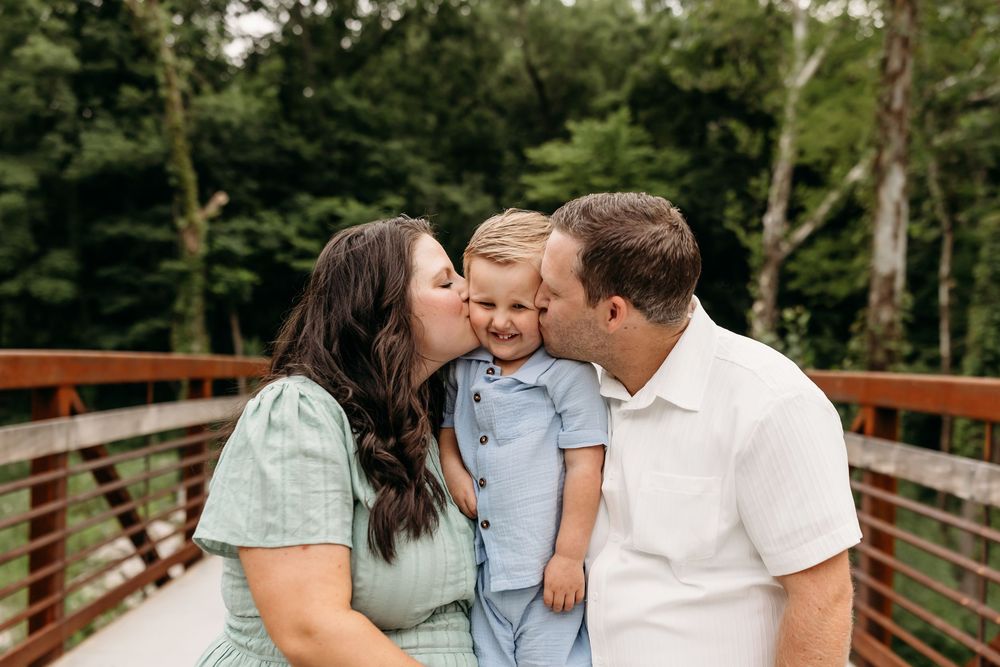 A couple kisses their smiling toddler on a wooden bridge surrounded by lush green trees.