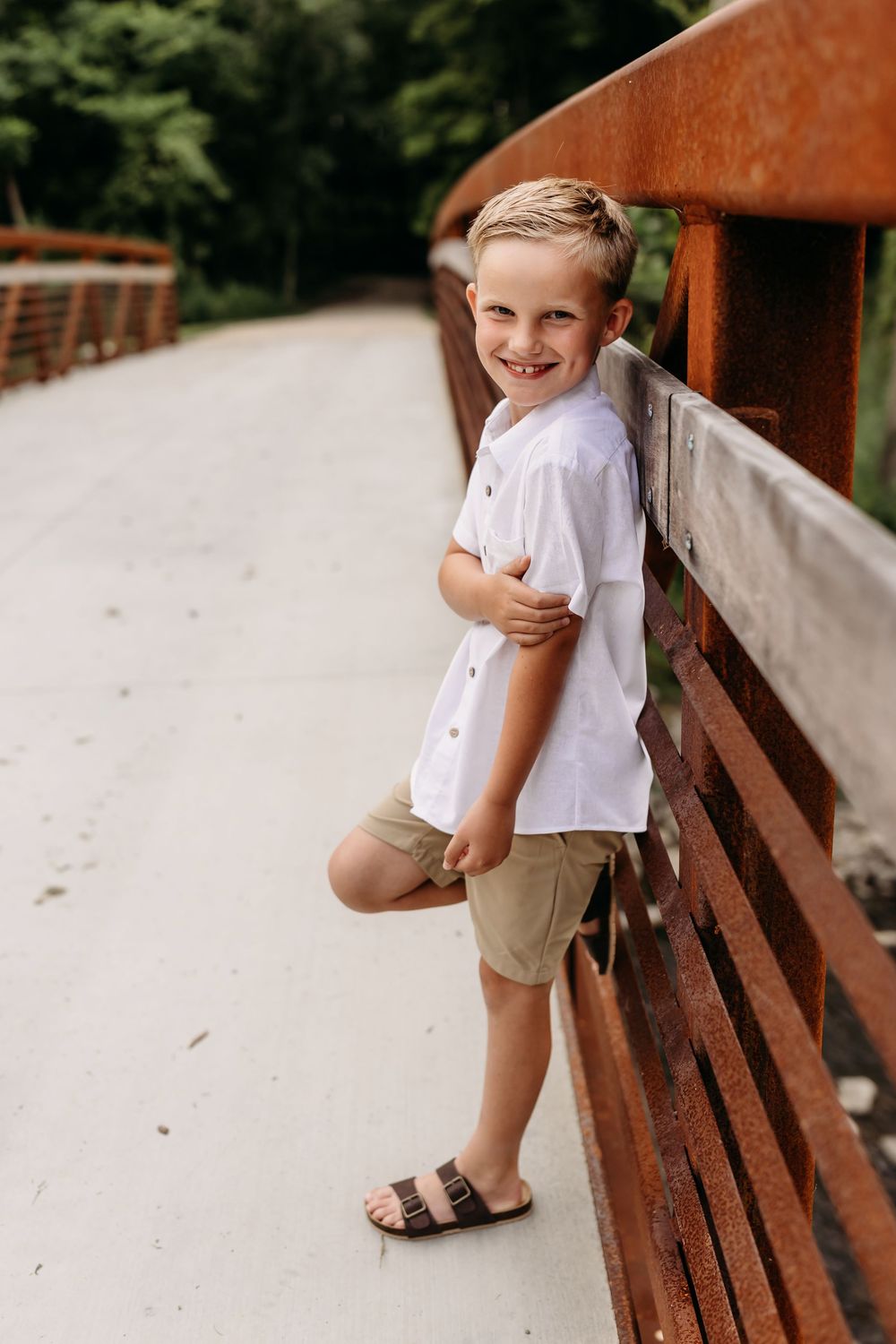 Smiling blonde boy in white shirt and khaki shorts leaning against a rustic bridge railing outdoors.