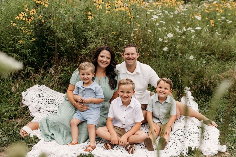 Family of five smiling together on a blanket in a wildflower meadow during a summer outdoor photo session.