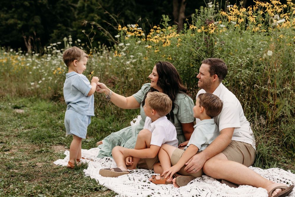 Family of five enjoying a picnic in a wildflower meadow, with mom reaching out to toddler son on a sunny day.
