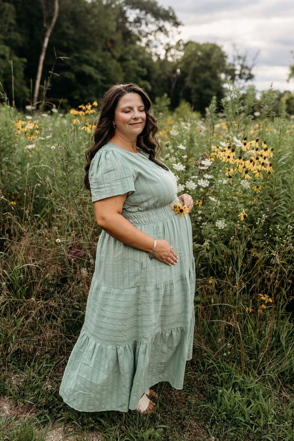 Woman in sage green midi dress standing in wildflower field with yellow and white blooms.