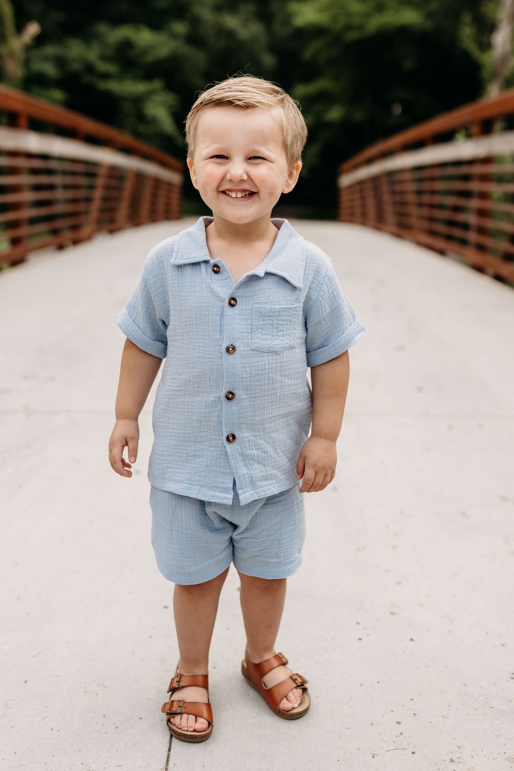 Smiling toddler boy in blue linen shirt and shorts with brown sandals standing on a bridge outdoors.