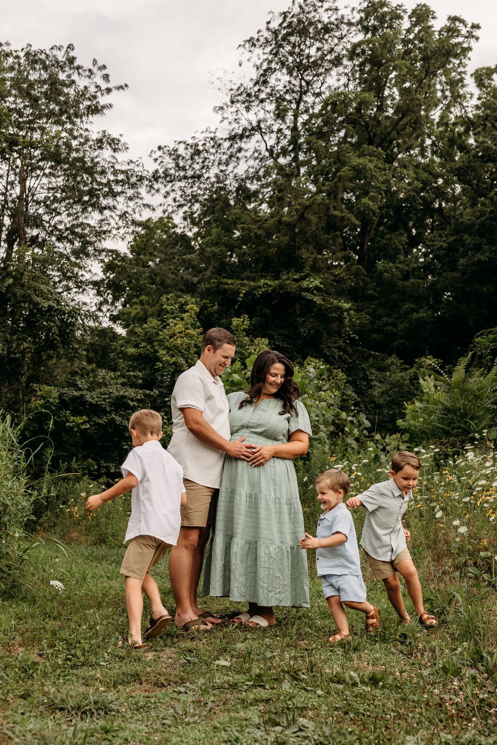 Pregnant woman in green dress with husband and two young boys in a lush outdoor field during a maternity photo session.