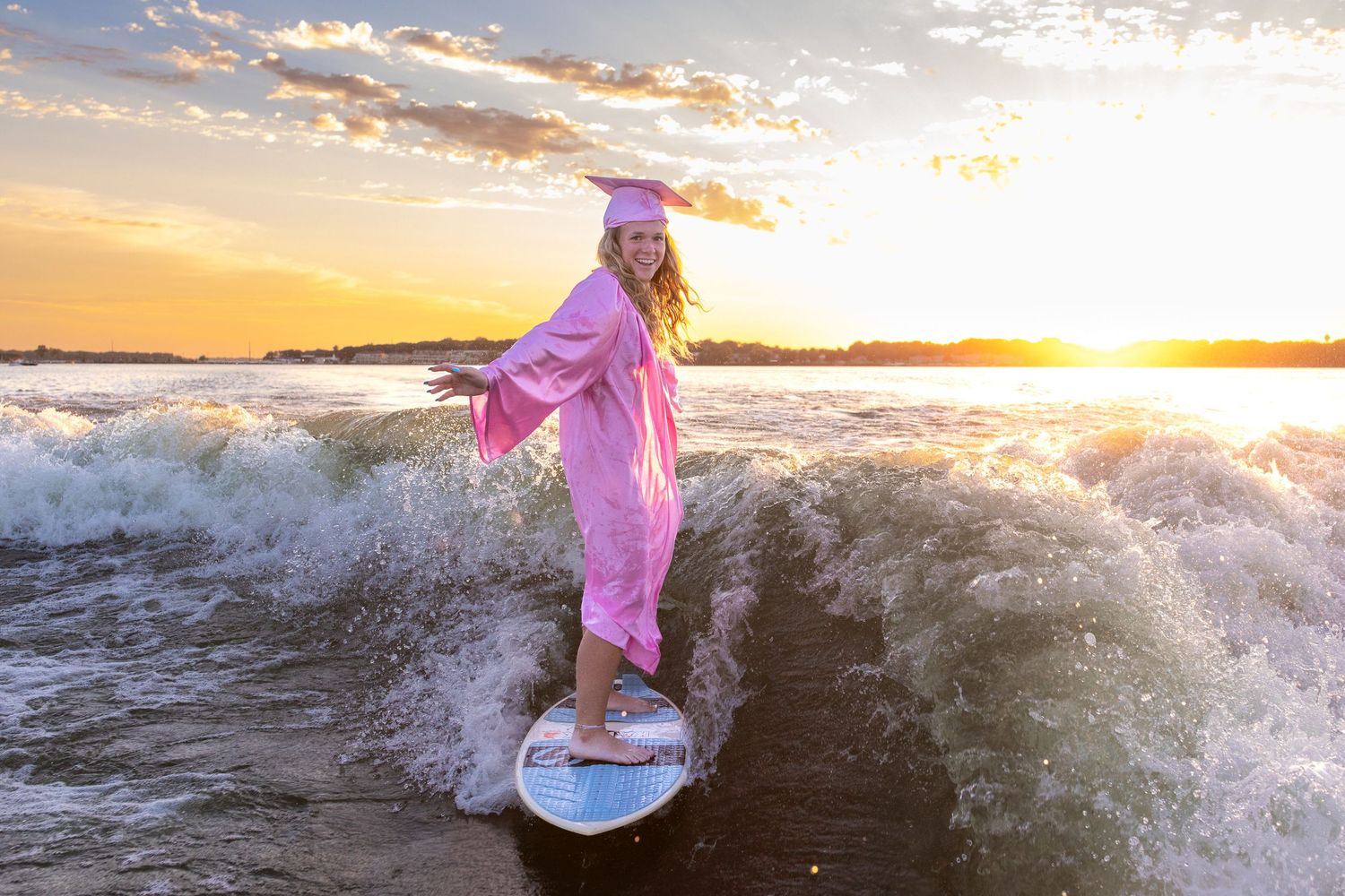 Senior girl in pink cap and gown rides a surfboard through crashing West Okoboji Lake waves at sunset.