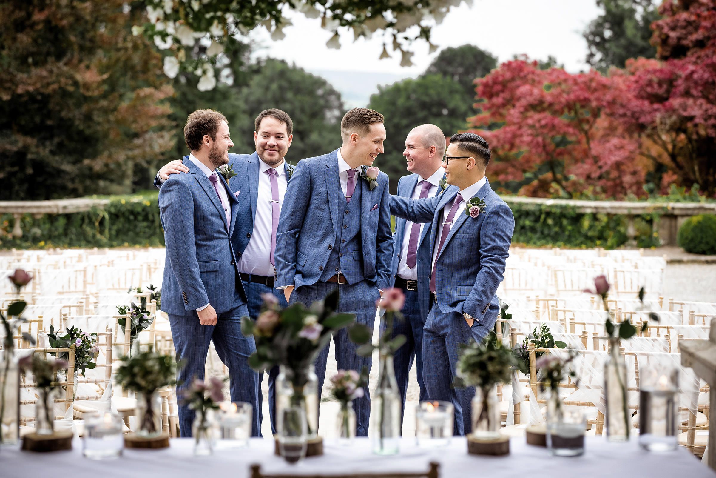 Five groomsmen in blue suits laughing together at an outdoor wedding venue with autumn trees and elegant table settings.