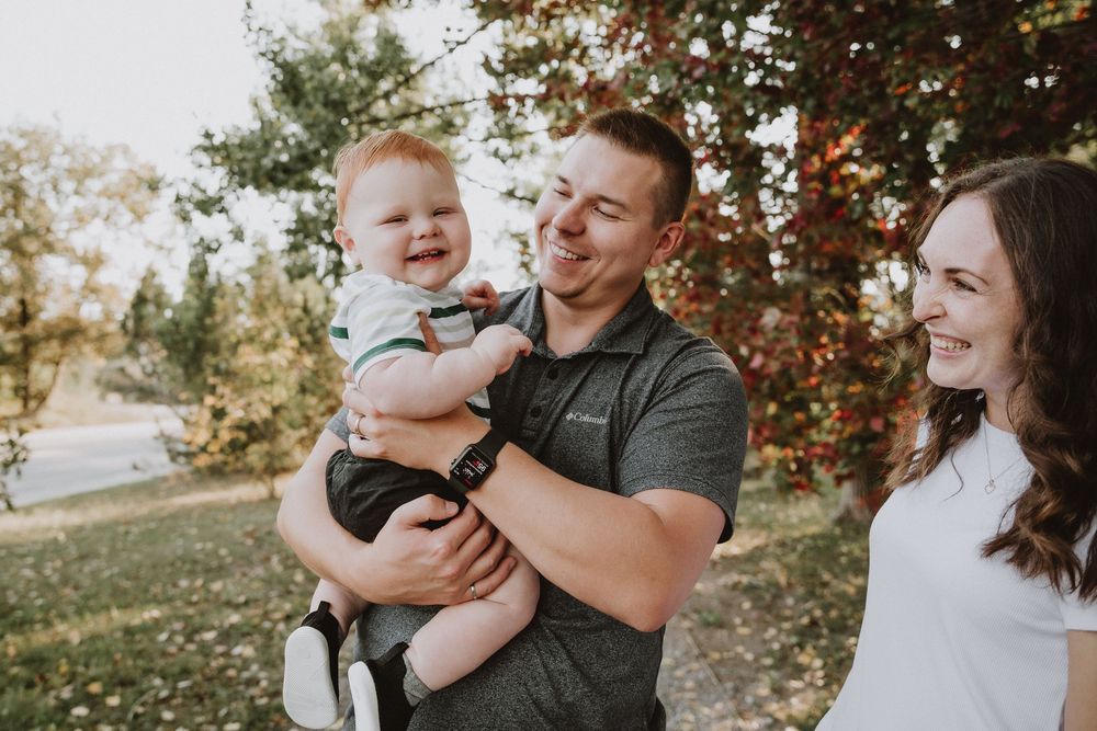 A young family smiles outdoors in autumn, father holding a happy baby while mother stands beside them near colorful trees.