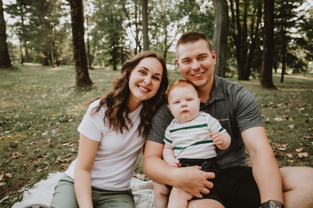 Young family with baby sitting on blanket in park, smiling outdoors among green trees.