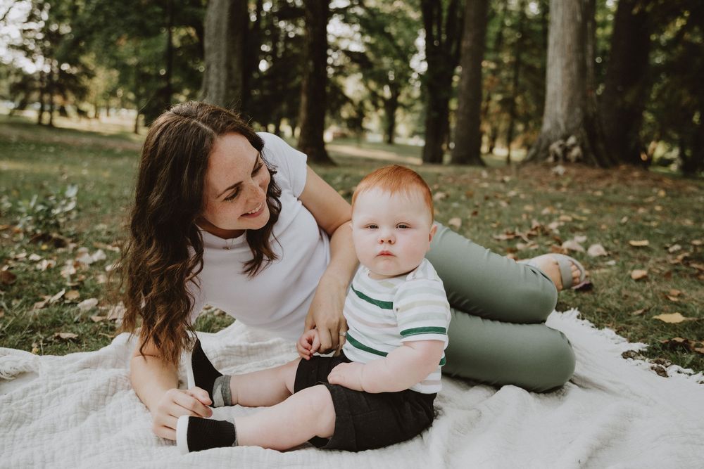 A young mother sits on a white blanket in a park with her red-haired baby boy on a sunny day.
