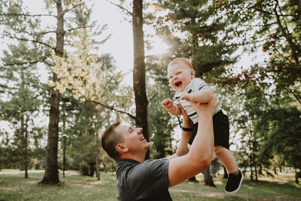 A joyful father lifts his laughing baby high in a sunny park, sharing a playful and loving outdoor moment.