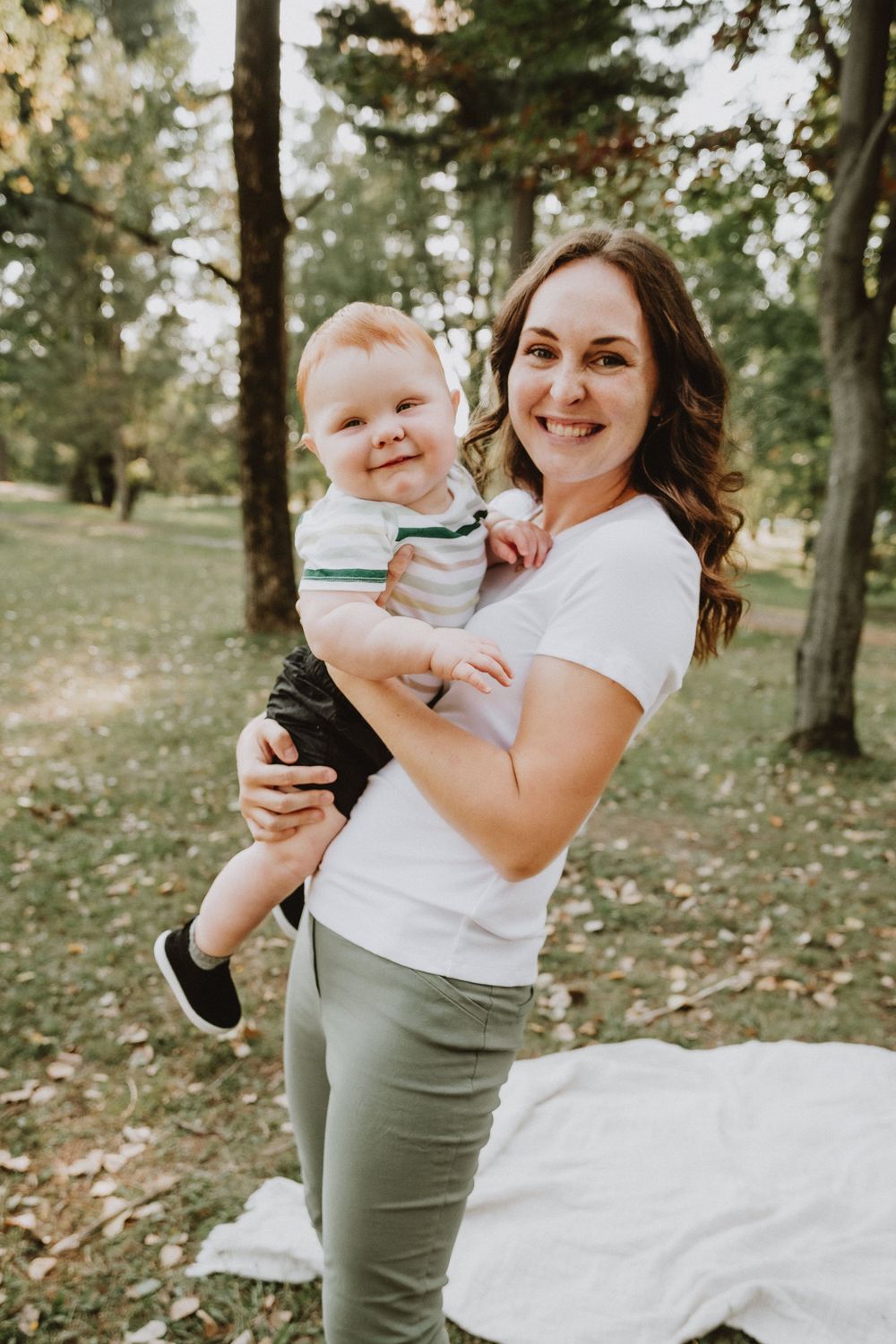 Smiling mother holding her happy baby boy outdoors in a lush green park setting.