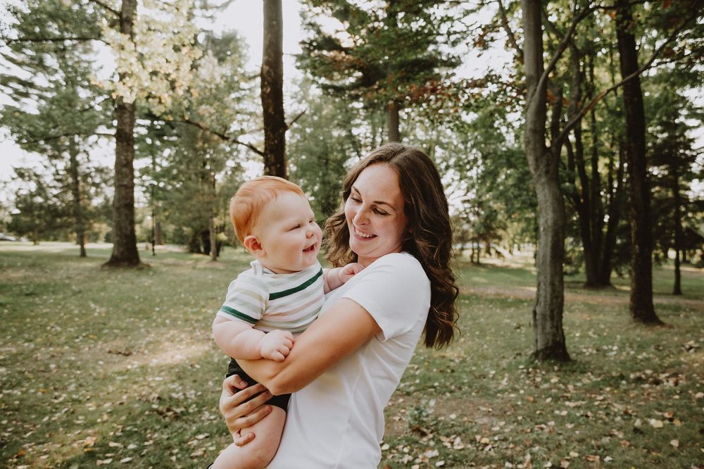 A young mother smiles while holding her happy red-haired toddler in a lush green park surrounded by tall trees.