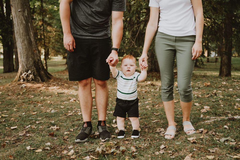 A baby taking first steps while holding hands with parents in a sunny park setting.